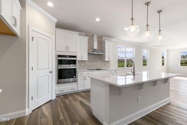 kitchen with wall chimney exhaust hood, double oven, decorative light fixtures, a center island with sink, and white cabinets