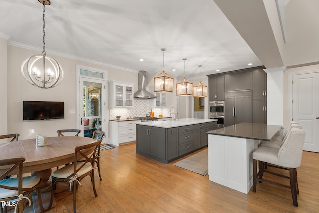 kitchen with a kitchen island with sink, stainless steel appliances, white cabinets, gray cabinetry, and wall chimney exhaust hood