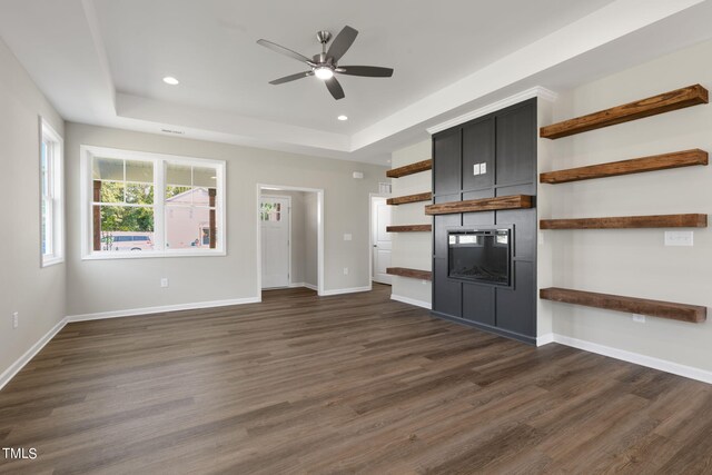 unfurnished living room with a fireplace, a tray ceiling, dark wood-type flooring, and ceiling fan