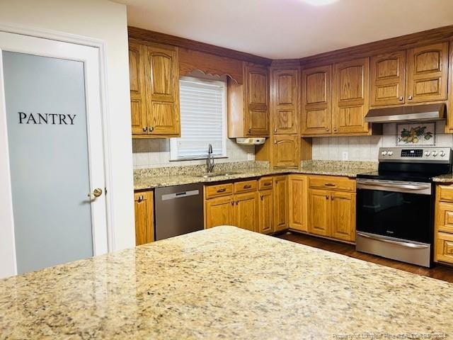 kitchen with stainless steel appliances, under cabinet range hood, light stone countertops, a sink, and backsplash
