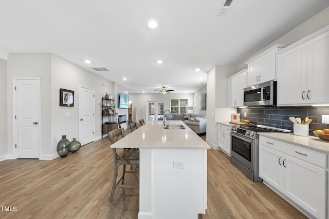 kitchen with light wood-type flooring, appliances with stainless steel finishes, an island with sink, decorative backsplash, and white cabinets