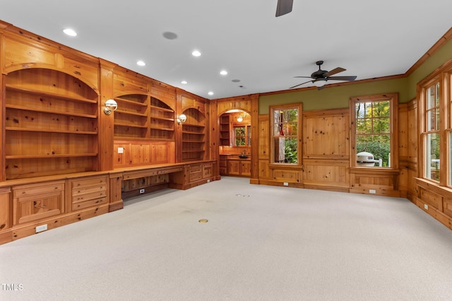 unfurnished living room with ceiling fan, light colored carpet, built in desk, and ornamental molding