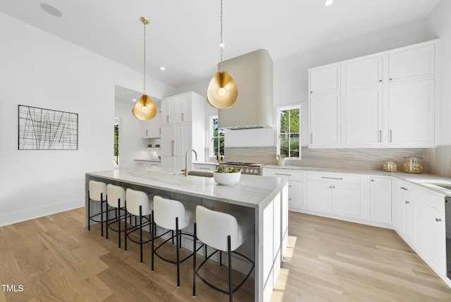 712 Daniels St, Raleigh, NC 27605 - kitchen featuring wall chimney range hood, a center island with sink, light wood-style flooring, a kitchen bar, and light stone countertops