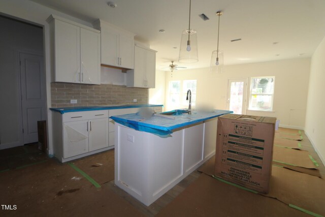 kitchen featuring backsplash, ceiling fan, white cabinets, hanging light fixtures, and an island with sink