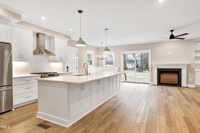 kitchen with wall chimney range hood, sink, appliances with stainless steel finishes, white cabinetry, and an island with sink