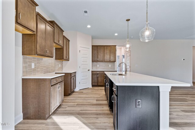 kitchen featuring backsplash, sink, decorative light fixtures, a center island with sink, and light hardwood / wood-style flooring