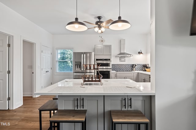 kitchen with kitchen peninsula, wall chimney range hood, gray cabinets, stainless steel appliances, and a kitchen bar