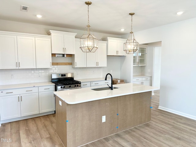 kitchen with stainless steel gas range oven, a kitchen island with sink, sink, decorative light fixtures, and white cabinetry