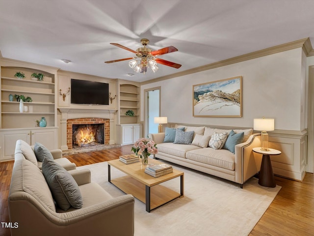 living room featuring a brick fireplace, ornamental molding, built in shelves, ceiling fan, and light hardwood / wood-style flooring