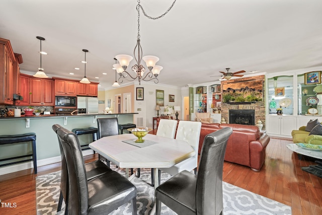 dining area featuring a stone fireplace, ornamental molding, dark wood-type flooring, and ceiling fan with notable chandelier