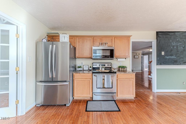 kitchen with light wood-type flooring, a textured ceiling, light brown cabinetry, appliances with stainless steel finishes, and stone countertops