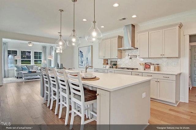 kitchen with ceiling fan, pendant lighting, a kitchen island with sink, wall chimney exhaust hood, and light hardwood / wood-style floors