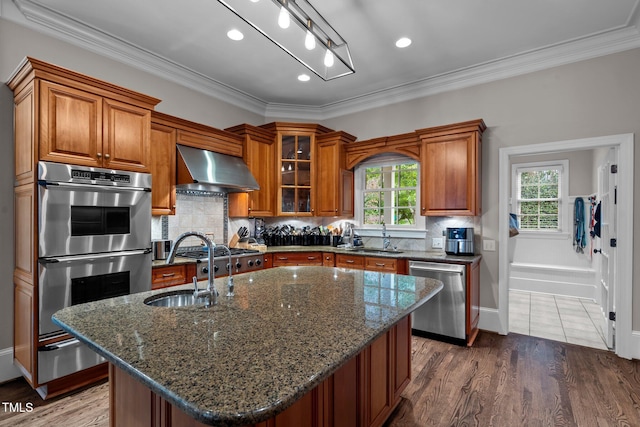 kitchen with a center island with sink, dark wood-type flooring, appliances with stainless steel finishes, and wall chimney range hood