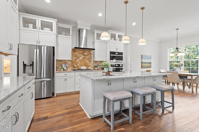 kitchen featuring wall chimney exhaust hood, an island with sink, stainless steel appliances, a breakfast bar, and pendant lighting