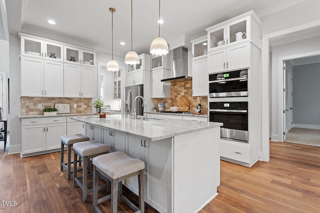 kitchen featuring white cabinetry, stainless steel appliances, wall chimney range hood, and a center island with sink