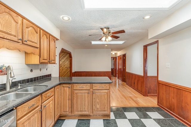 kitchen featuring a wainscoted wall, stainless steel dishwasher, a sink, a skylight, and a peninsula