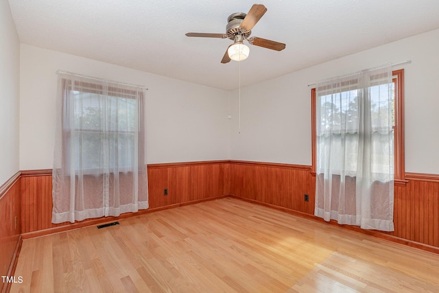 spare room with a wainscoted wall, light wood-type flooring, visible vents, a ceiling fan, and wooden walls
