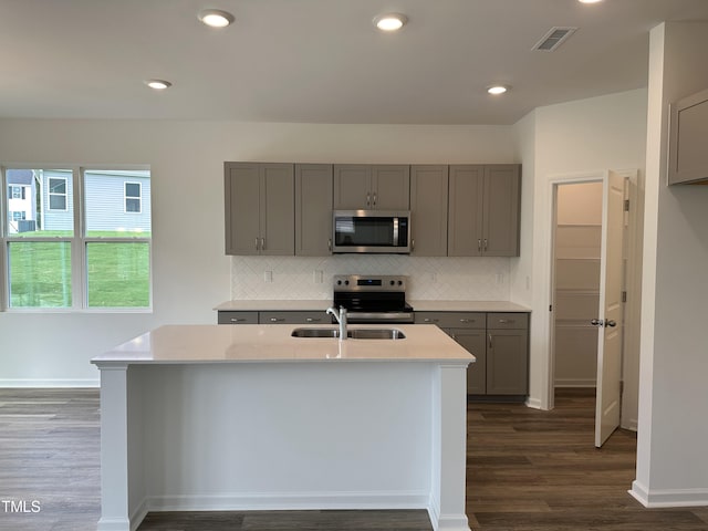 kitchen with gray cabinetry, sink, an island with sink, stainless steel appliances, and dark wood-type flooring