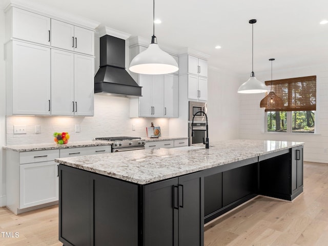 kitchen with a large island with sink, custom range hood, decorative light fixtures, light hardwood / wood-style floors, and white cabinetry