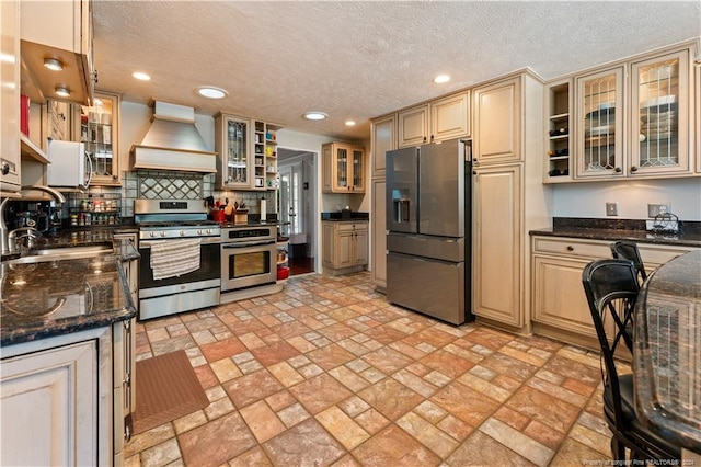 kitchen featuring custom exhaust hood, hanging light fixtures, backsplash, stainless steel appliances, and dark stone countertops