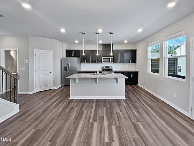 kitchen featuring appliances with stainless steel finishes, pendant lighting, dark wood-type flooring, light stone counters, and a kitchen island with sink