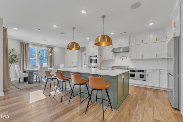 kitchen featuring white cabinets, light countertops, freestanding refrigerator, under cabinet range hood, and an island with sink