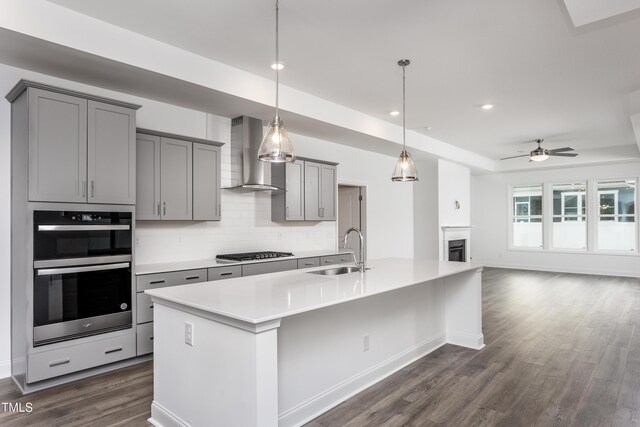 kitchen featuring gray cabinetry, wall chimney exhaust hood, sink, decorative light fixtures, and a center island with sink