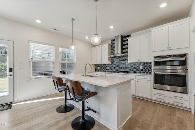 kitchen featuring hanging light fixtures, double oven, white cabinets, and wall chimney range hood