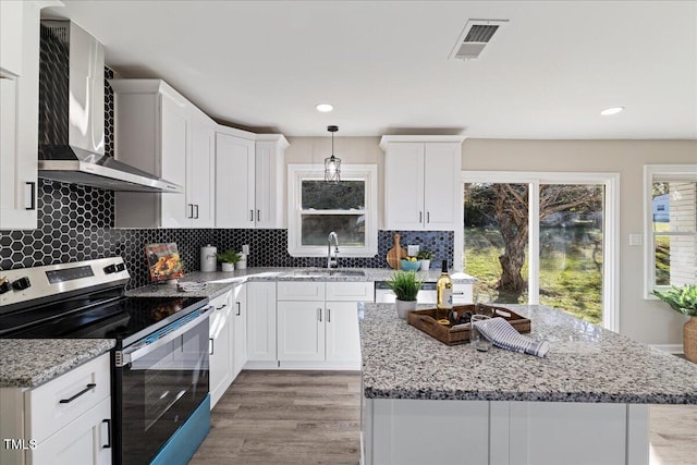 kitchen featuring wall chimney range hood, light wood-type flooring, hanging light fixtures, stainless steel electric range, and white cabinets