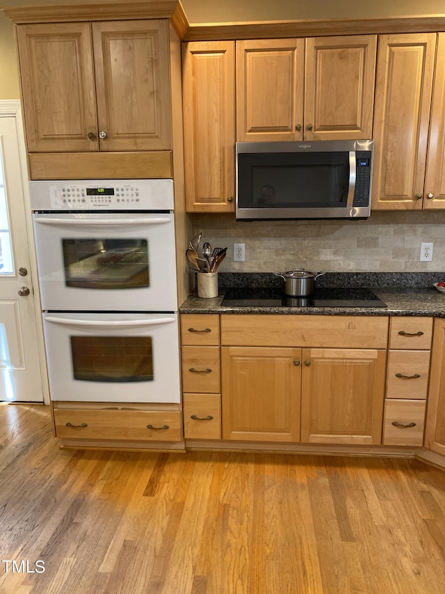 kitchen with black electric stovetop, tasteful backsplash, double oven, dark stone counters, and light hardwood / wood-style flooring