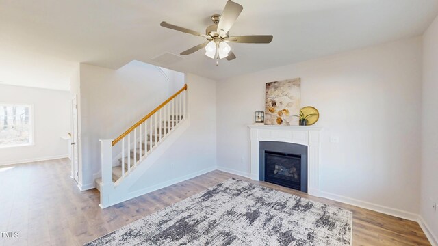unfurnished living room with ceiling fan, a glass covered fireplace, wood finished floors, stairs, and baseboards