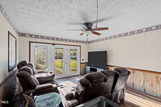 living room with french doors, ceiling fan, light wood-type flooring, a textured ceiling, and wood walls