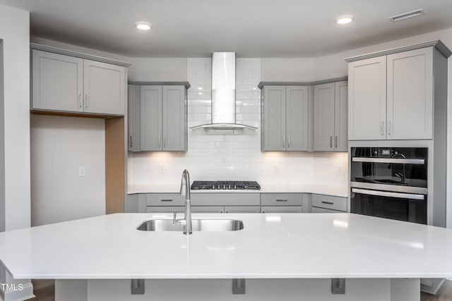 kitchen with sink, gray cabinets, a kitchen island with sink, and wall chimney range hood