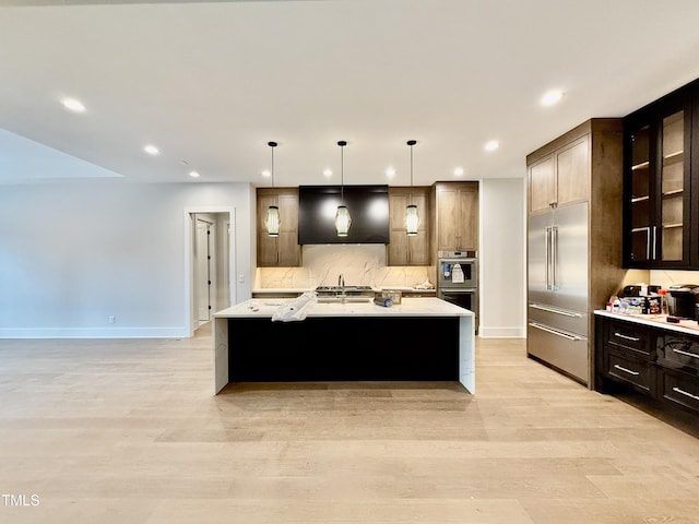 kitchen featuring hanging light fixtures, light wood-type flooring, a center island with sink, stainless steel appliances, and backsplash