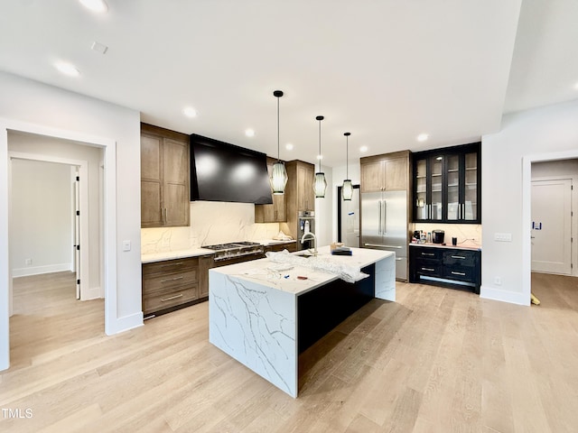 kitchen with a kitchen island with sink, stainless steel appliances, light stone countertops, decorative light fixtures, and custom exhaust hood