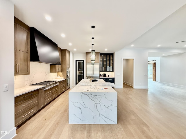 kitchen featuring wall chimney range hood, stainless steel appliances, tasteful backsplash, a center island with sink, and decorative light fixtures