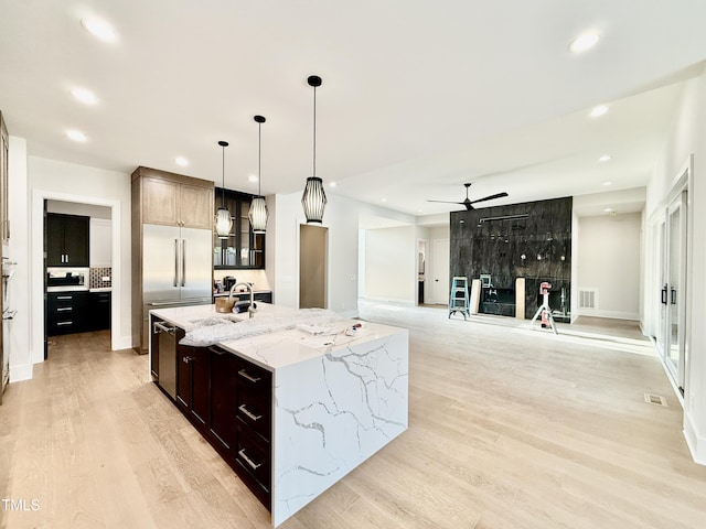 kitchen with a large island with sink, hanging light fixtures, stainless steel built in fridge, light hardwood / wood-style floors, and light stone countertops