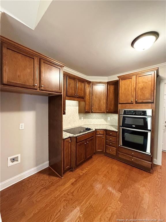 kitchen with light stone countertops, decorative backsplash, black electric stovetop, double oven, and light hardwood / wood-style flooring