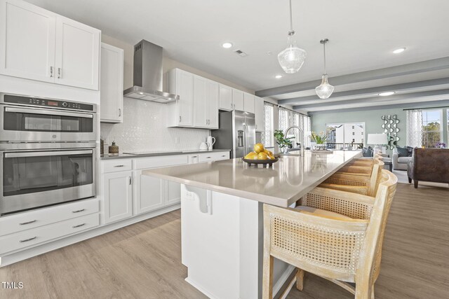 kitchen with wall chimney range hood, light hardwood / wood-style flooring, appliances with stainless steel finishes, decorative light fixtures, and a kitchen bar