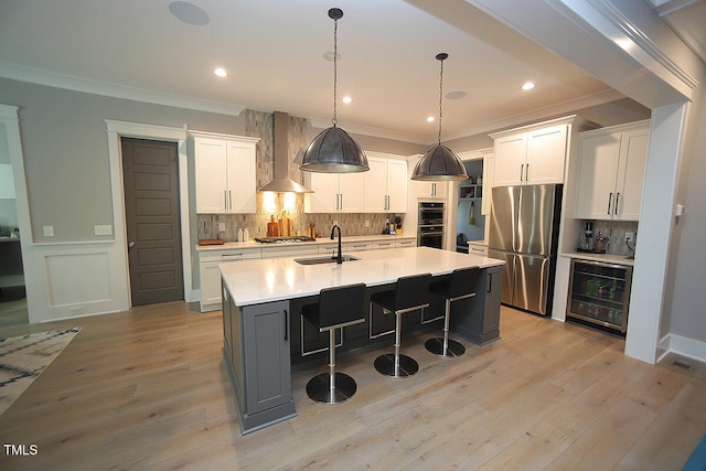 kitchen with wall chimney range hood, white cabinetry, stainless steel appliances, and an island with sink