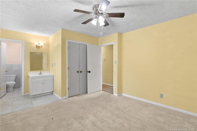 unfurnished bedroom featuring a closet, ensuite bath, light colored carpet, a textured ceiling, and ceiling fan