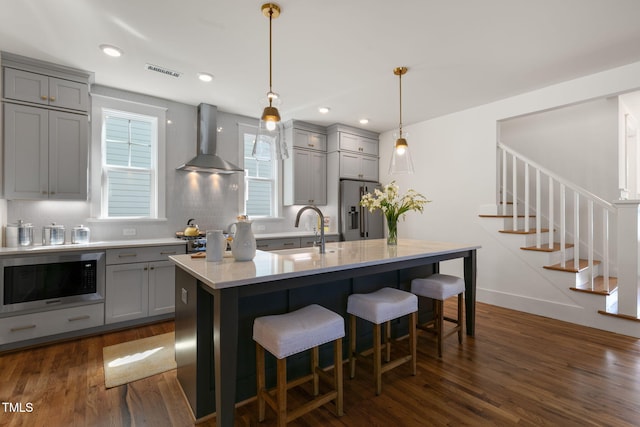 kitchen featuring wall chimney range hood, an island with sink, dark hardwood / wood-style flooring, stainless steel appliances, and pendant lighting