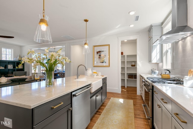 kitchen featuring appliances with stainless steel finishes, sink, wall chimney exhaust hood, light hardwood / wood-style flooring, and a center island with sink