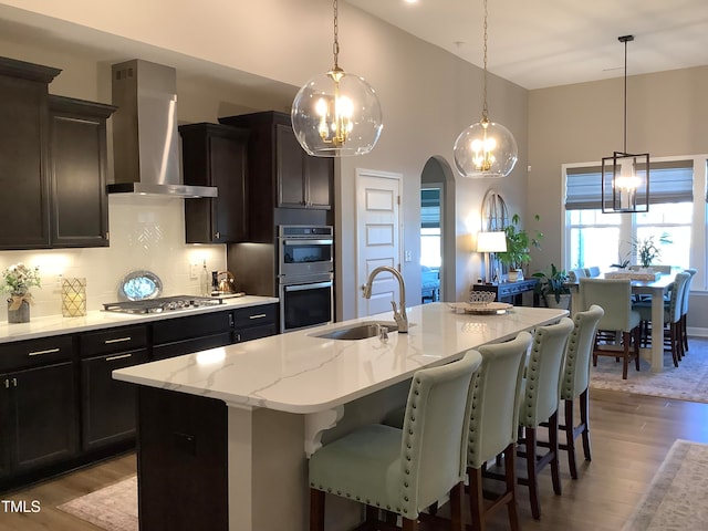 kitchen featuring wall chimney range hood, a center island with sink, appliances with stainless steel finishes, light wood-type flooring, and sink