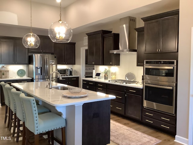 kitchen with wall chimney range hood, a kitchen island with sink, sink, pendant lighting, and stainless steel appliances