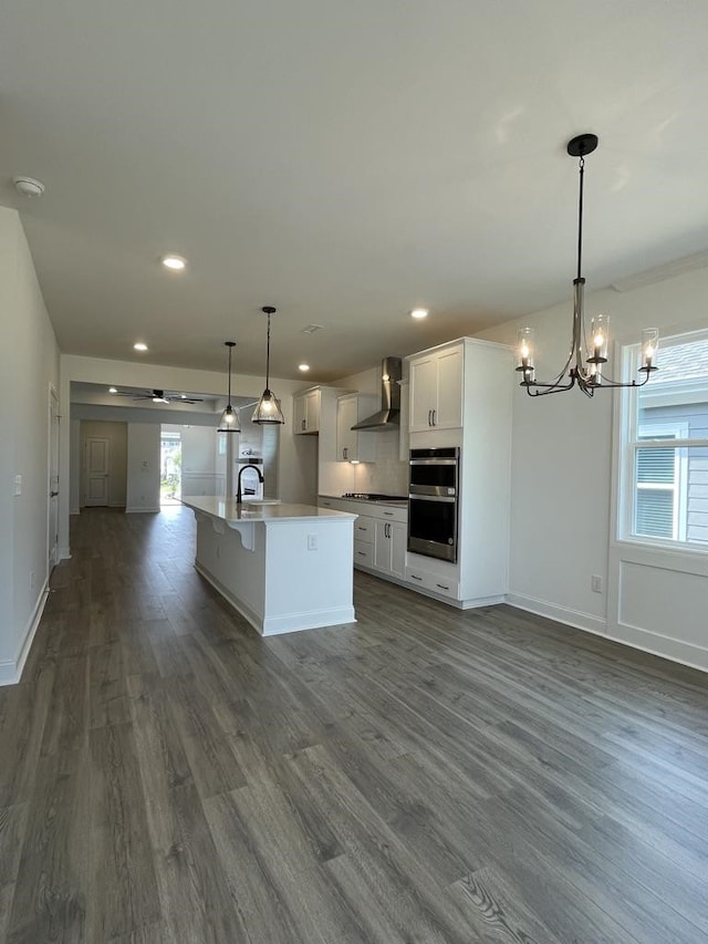 kitchen with wall chimney range hood, appliances with stainless steel finishes, an island with sink, hanging light fixtures, and dark wood-type flooring