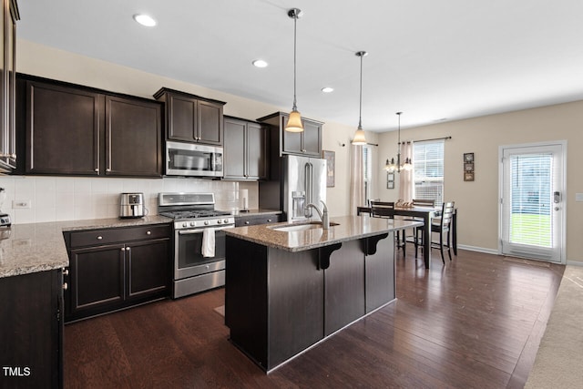 kitchen with a center island with sink, appliances with stainless steel finishes, light stone countertops, dark hardwood / wood-style floors, and decorative light fixtures