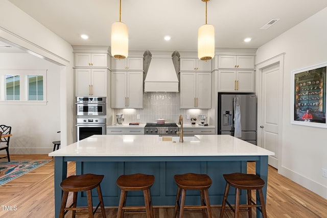 kitchen with an island with sink, hanging light fixtures, custom range hood, sink, and appliances with stainless steel finishes