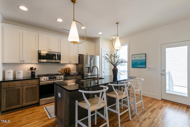 kitchen featuring white cabinets, a center island with sink, appliances with stainless steel finishes, light wood-type flooring, and decorative light fixtures