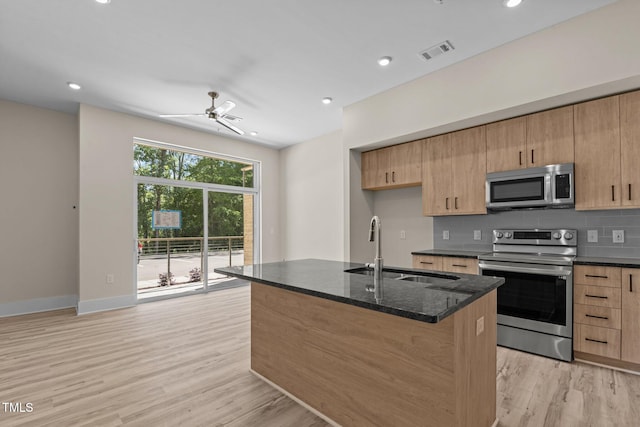 kitchen featuring appliances with stainless steel finishes, a kitchen island with sink, dark stone counters, light wood-type flooring, and sink